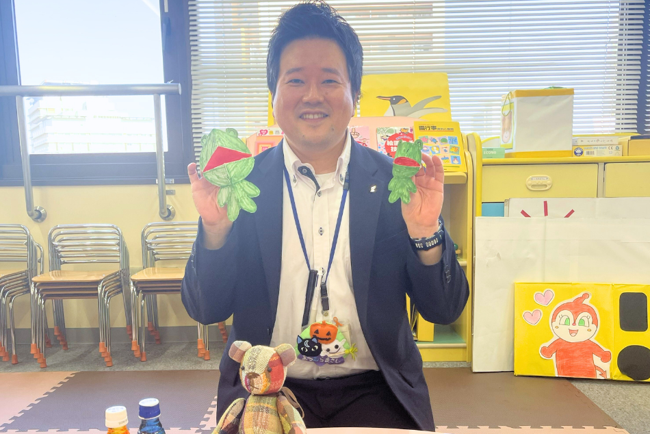 手作りカエルの玩具を両手で持って笑みを浮かべている松村先生の写真